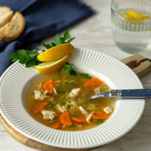 Hausgemachte Hühnerbrühe in einem weißen tiefen Teller mit orangefarbenen Möhrenscheiben, Hühnerfleisch, Nudeln, Petersilie auf dem Tellerrand liegen Zitronenschnitze