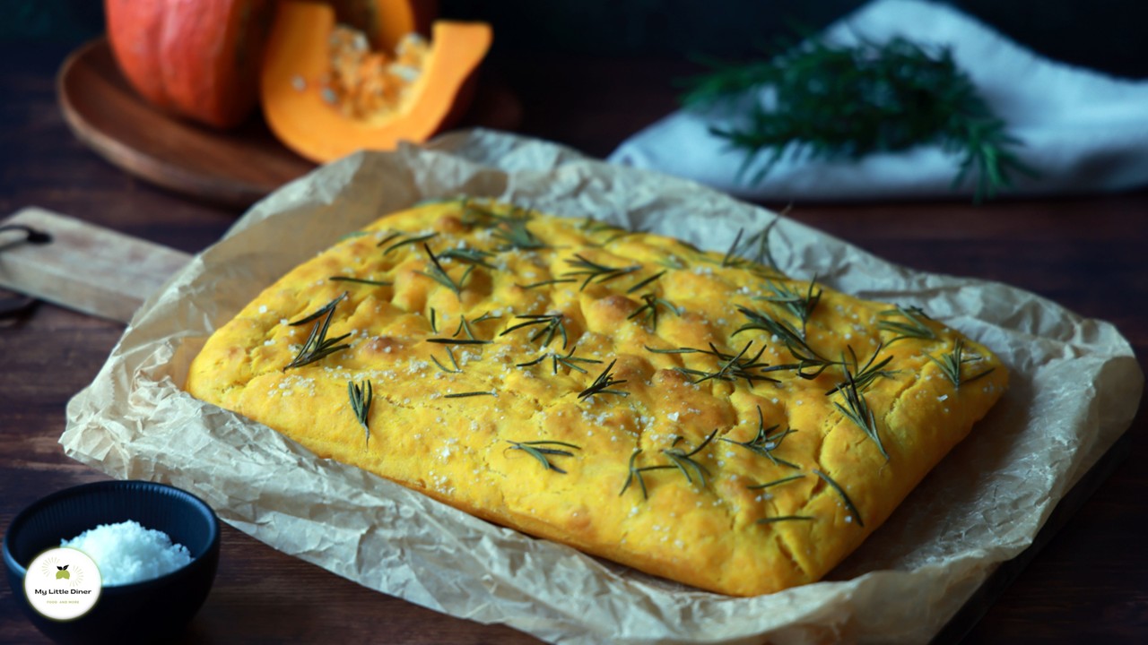 Eine frisch gebackene gelbe Kürbis Focaccia liegt noch auf Backpapier auf einem Holzbrett. Die Oberfläche ist mit Rosmarinnadeln und Meersalzflocken bestreut.