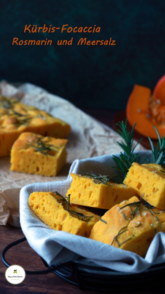Goldgelbe Stücke von einer Kürbis Focaccia liegen in einem Brotkorb auf einem Holztisch. Dahinter steht noch das Backblech mit der restlichen Focaccia. Im Hintergrund ein angeschnittener Kürbis.