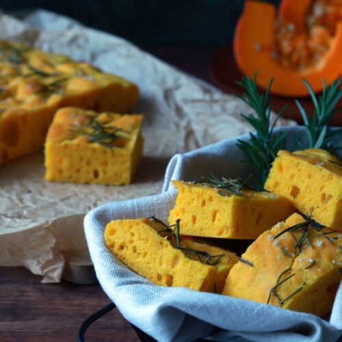 Sonnengelbe Quadrate von luftig gebackener Kürbis Focaccia liegen in einem Brotkorb auf einem dunklen Holztisch.