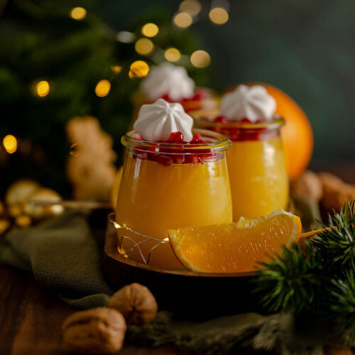 Drei Dessertgläser auf einem Holzbrett, gefüllt mit erfrischendem Orangengelee, getoppt mit roten Granatapfelkernen und weißen Baiser Tupfen, weihnachtlich dekoriert