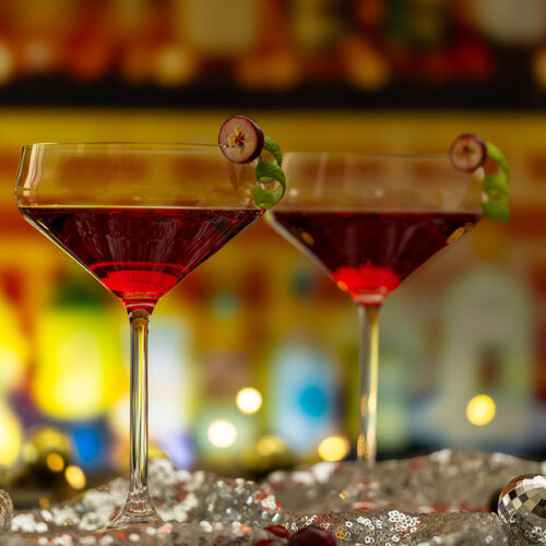 Zwei Cocktailgläser mit rotem Getränk stehen auf einem Bartresen und werden von hinten angeleuchtet