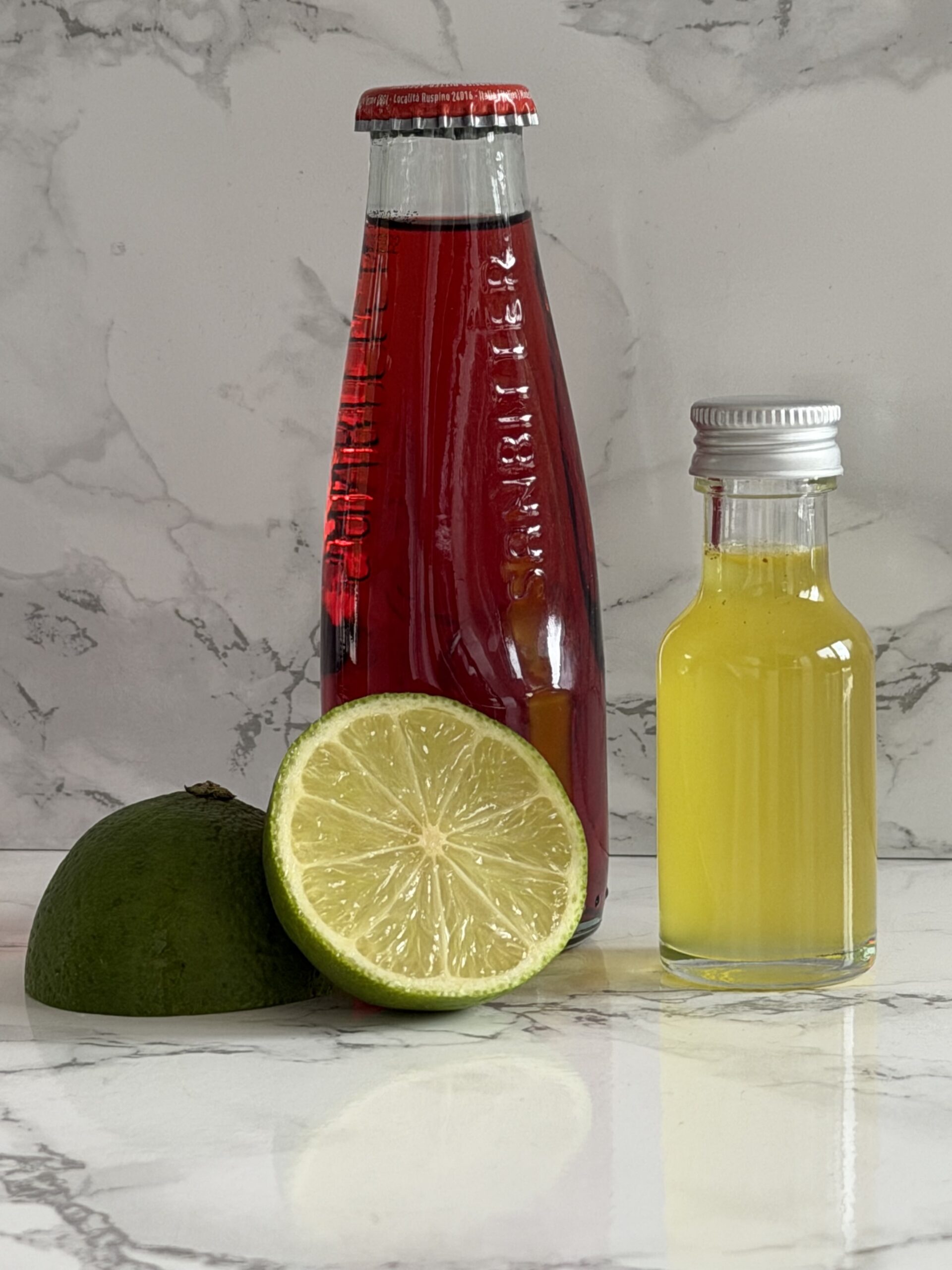 Auf einer weißen Marmorfläche liegt eine aufgeschnittene Limette neben einer Flasche mit rotem Sanbitter und einer kleinen Flaschen selbstgemachtem Orangen-Öl-Zucker