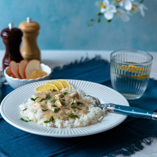 Ein weißer Teller steht auf einer blauen Stoffserviette. Hühner-Ragout in cremiger heller Sauce liegt auf einer Portion Reis, Ringe von grünen Frühlingszwiebeln als Dekoration sind darüber gestreut.